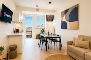 A modern living space with a dining area, a kitchen with a visible sink, a flat-screen TV, and a large window offering a city view. - DomusNova Finestra Penthouse historic center Turin (Torino)