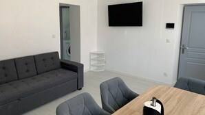 Uma sala de estar moderna com um sofá cinza, uma mesa de jantar de madeira com cadeiras, uma TV de tela plana na parede e uma porta com puxador.
