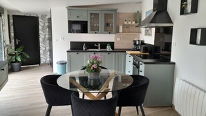 A modern kitchen with grey cabinets, a black range hood, and a round glass table with two black chairs. - Accommodation with a landscaped patio with trees. Ideal for a moment for two. (Ailly-sur-Noye)