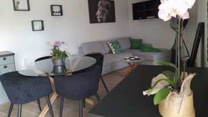 A modern living room with a grey sofa, a coffee table, and a dining area with two chairs. There are framed pictures on the wall and a potted plant on the table. - Accommodation with a landscaped patio with trees. Ideal for a moment for two. (Ailly-sur-Noye)