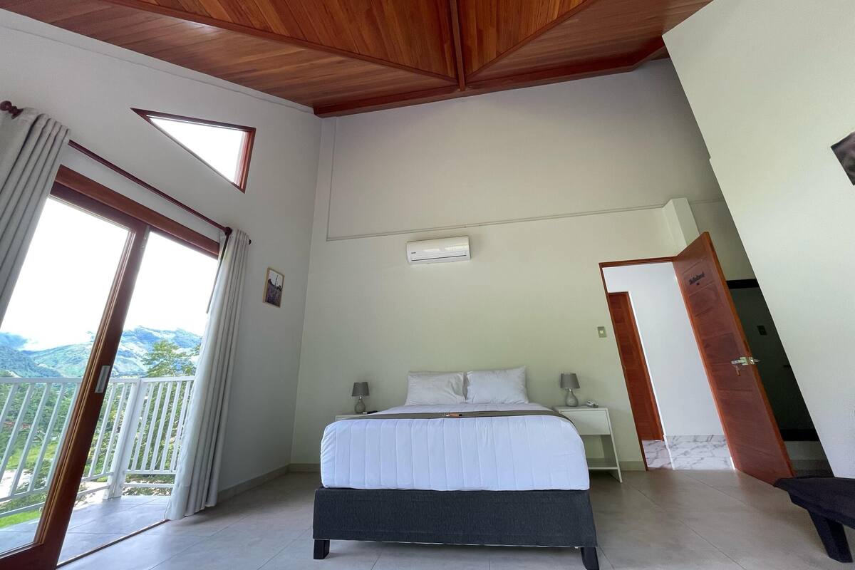 Een slaapkamer met een bed, nachtkastjes, een airconditioner, een balkon met gordijnen en een houten plafond.