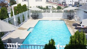 Une piscine rectangulaire entourée d’une clôture blanche, un espace patio avec des chaises et des parasols, et une vue sur les maisons voisines.