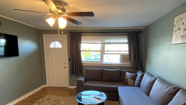 Un salon avec un ventilateur de plafond, une table basse en verre, un canapé marron et une porte.