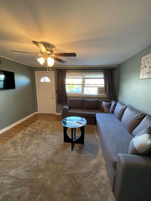 A living room with a ceiling fan, a glass coffee table, a brown sofa, and a door. - 23 B ST Apartments  (Hampton)