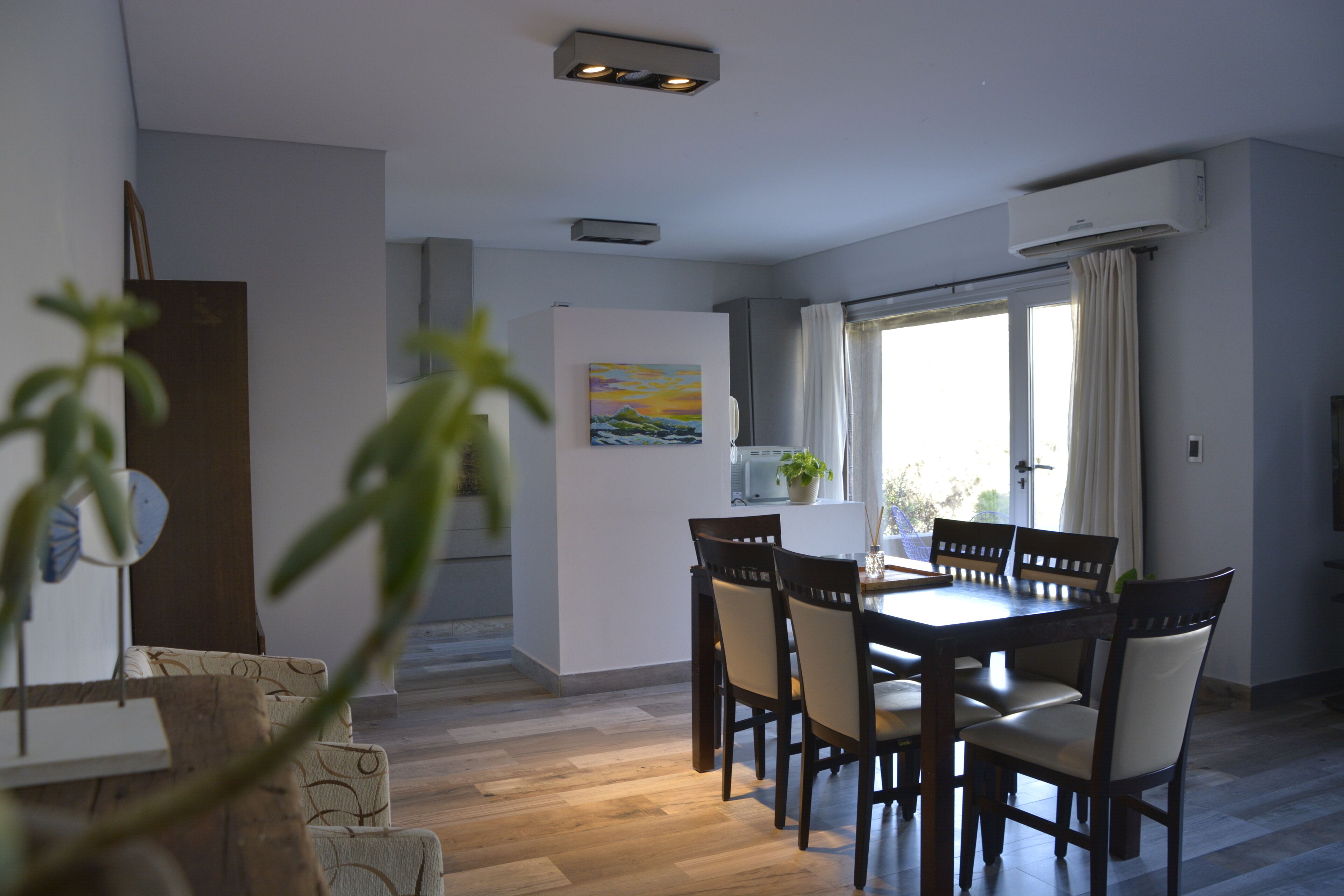 Uma sala de jantar com uma mesa e cadeiras de madeira, um quadro na parede e uma janela com cortinas.