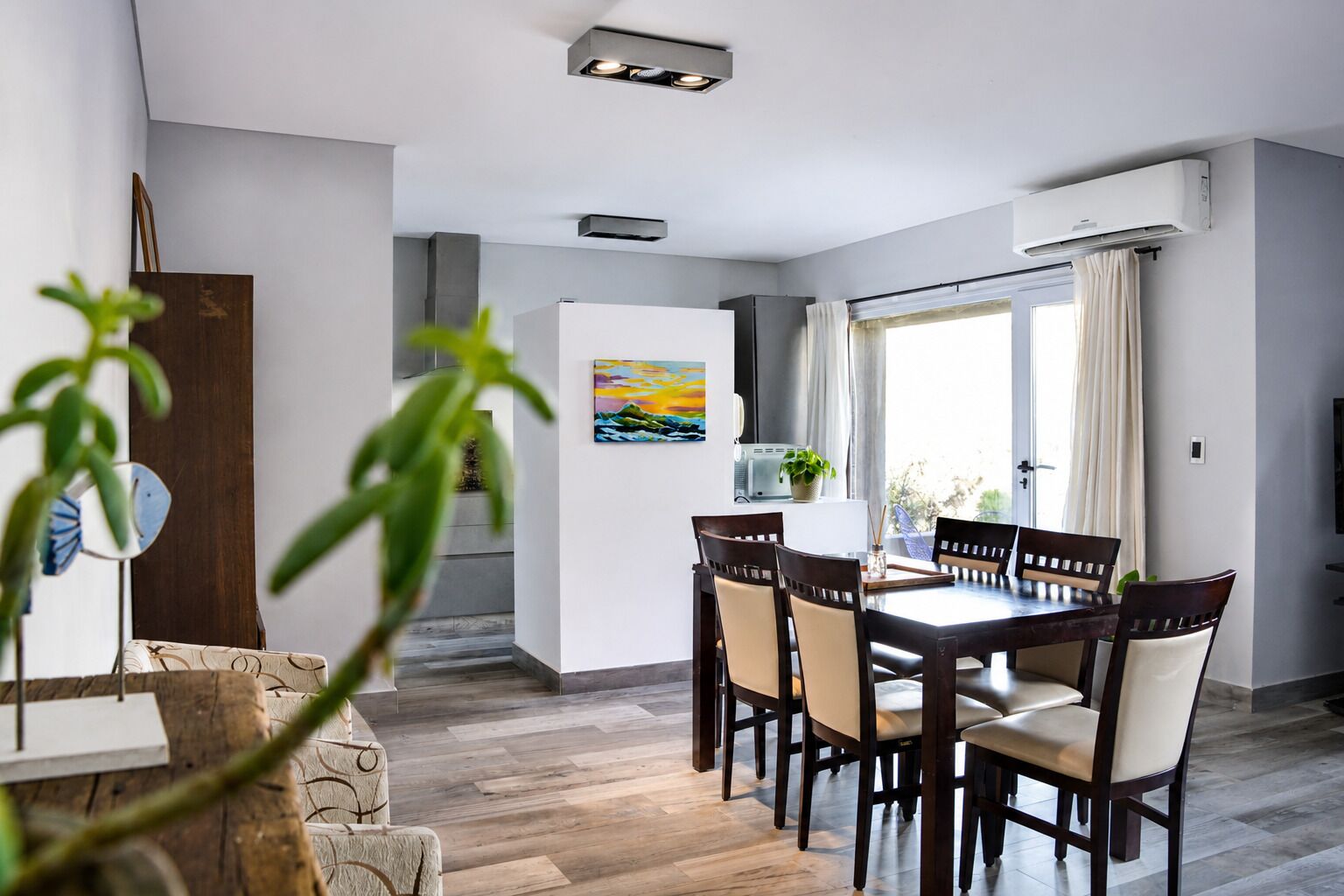 A dining area with a table and chairs, a painting on the wall, and an air conditioning unit.