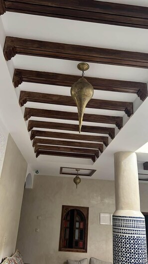 A room with exposed wooden beams, a decorative brass ceiling medallion, a traditional arched window, and a patterned column. - RIAD RKIYA (Tiznit)