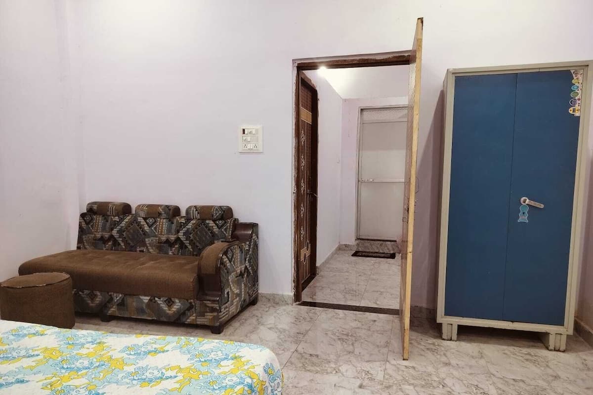 Une pièce avec un canapé à motifs, un lit recouvert d’une couvre-lit à fleurs, une armoire bleue et une porte donnant sur une autre pièce.