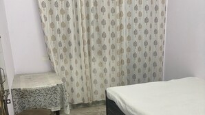 A bedroom with a bed, a small table, two ottomans, a curtain, and a wall-mounted light fixture. - Hotel Gayatri Palace (Chandauli)
