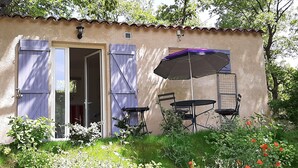 A small building with a door and windows, a patio with a table and chairs, and a garden with flowers and plants. - Country cottage, calm and relaxation in the sun. (Sainte-Croix-à-Lauze)