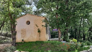 A small, rustic building with a circular window and lattice patterns on its walls, surrounded by lush greenery and a variety of plants. - Country cottage, calm and relaxation in the sun. (Sainte-Croix-à-Lauze)