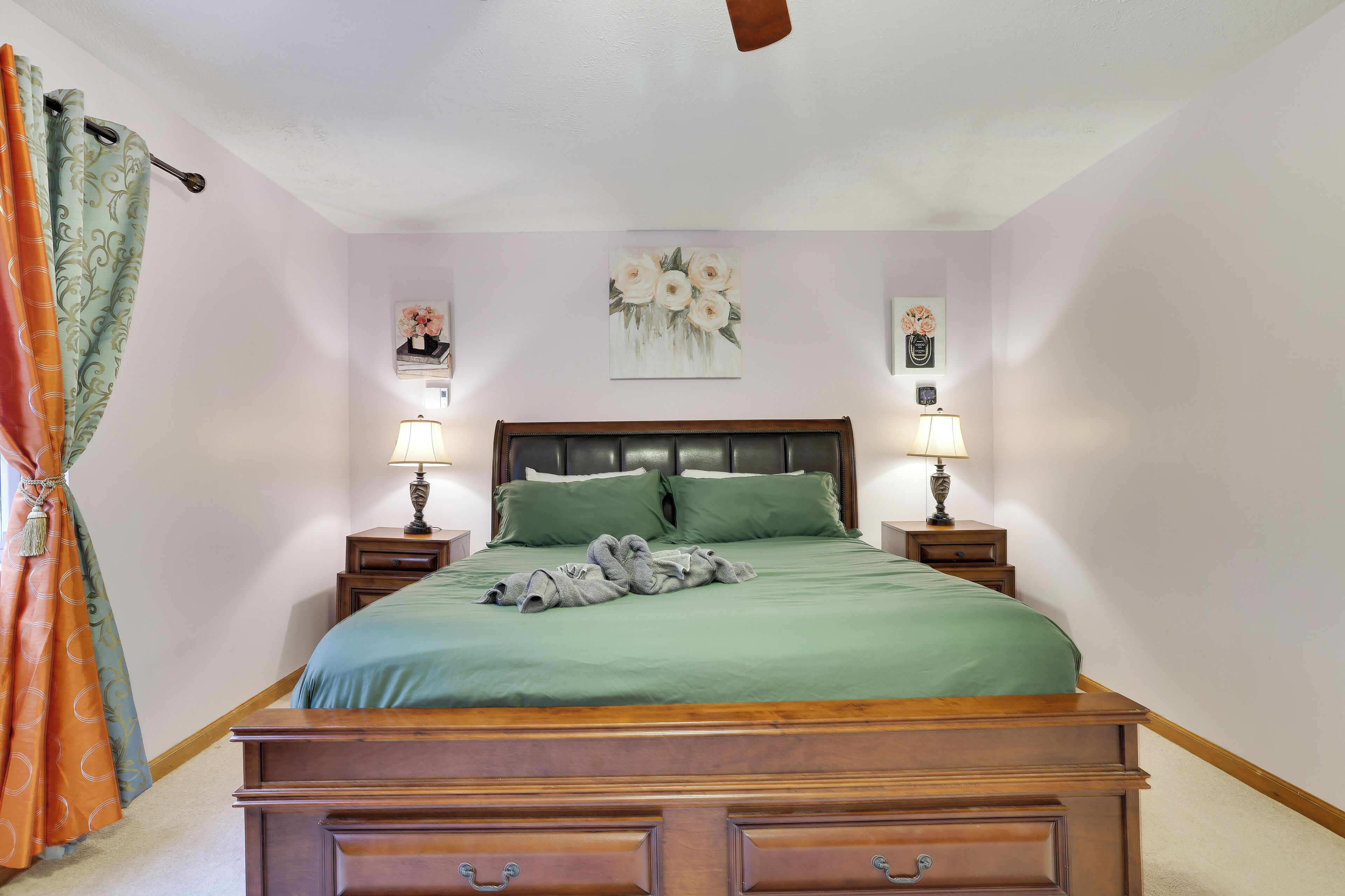 A bedroom with a wooden bed, green bedding, two bedside lamps, a headboard, and a floral artwork on the wall.