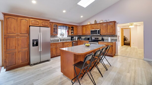 A kitchen with wooden cabinets, stainless steel appliances, a central island with chairs, and a ceiling light. - SPACIOUS 6-bedroom house with TWO MASTER BR AND BA, GAME ROOM, HOT TUB (Long Pond)