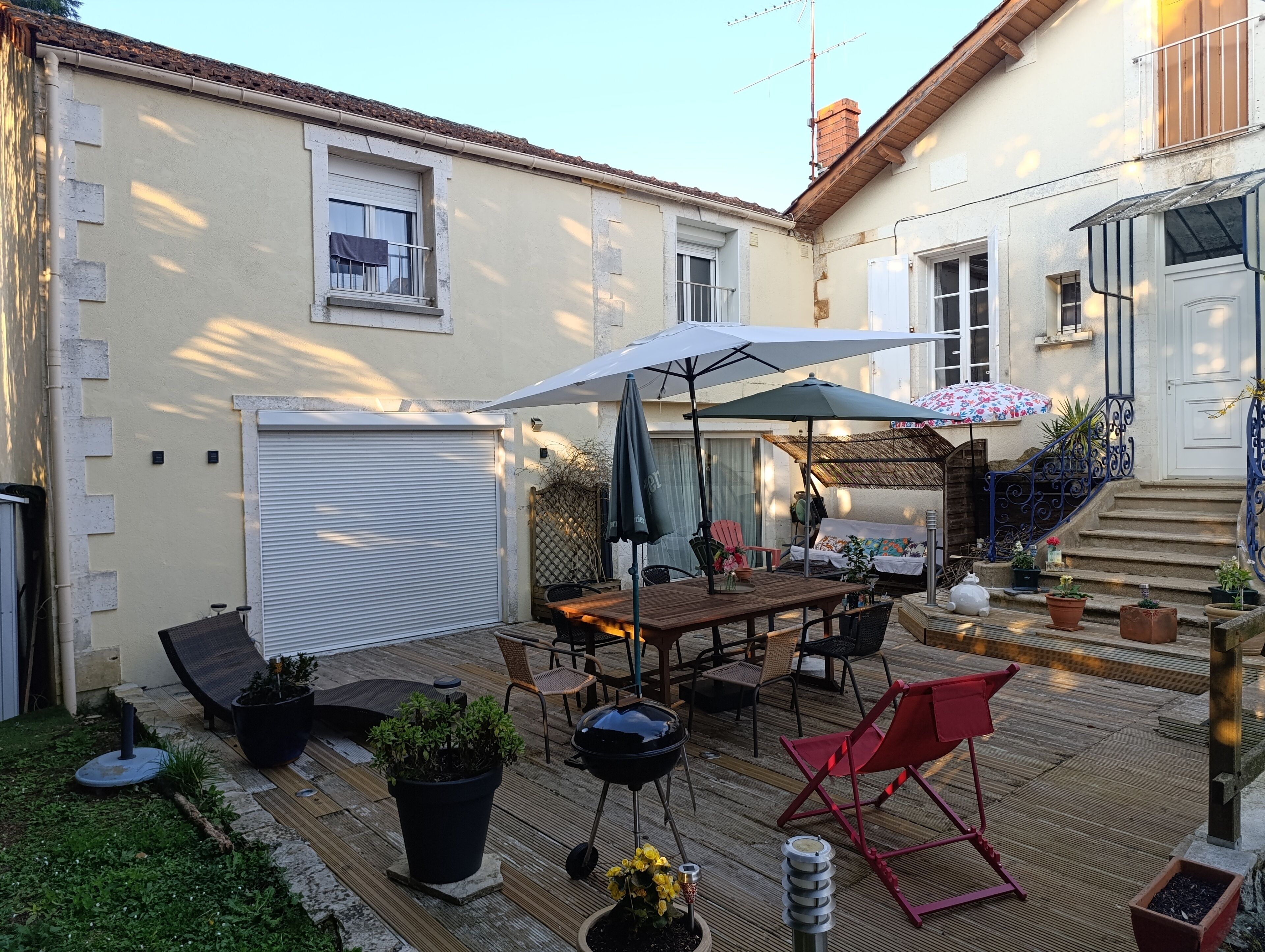 Un patio con mesa de madera, sillas y una parrilla. Hay plantas en macetas y una silla plegable roja.