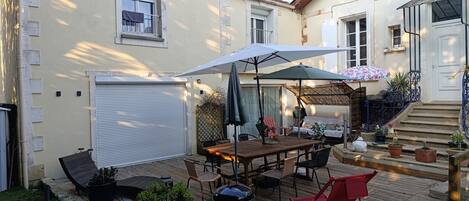 Un patio con mesa de madera, sillas y una parrilla. Hay plantas en macetas y una silla plegable roja.