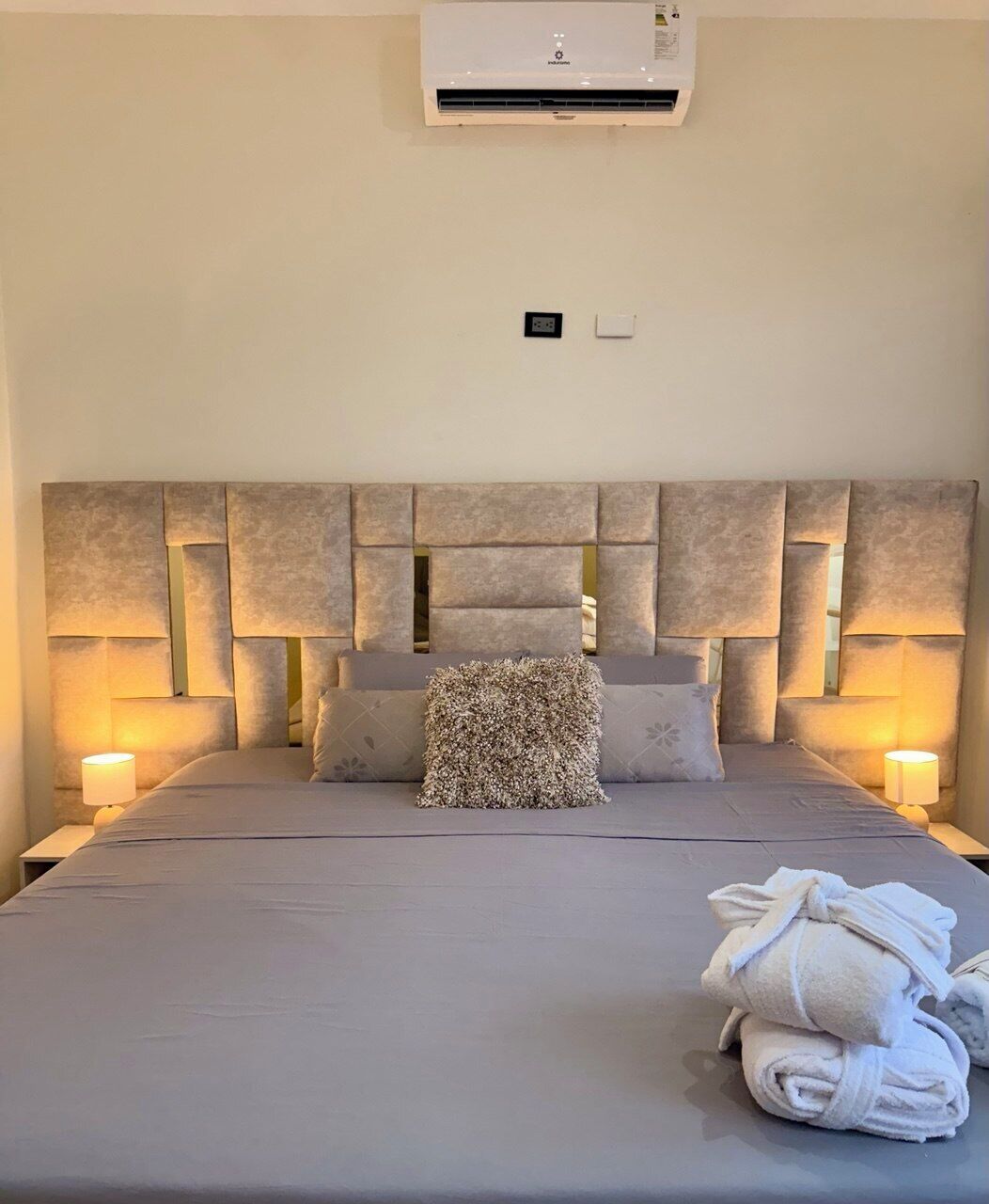Una camera d'albergo con un letto grande, una testiera geometrica, due lampade ai comodini, un condizionatore d'aria e un asciugamano arrotolato sul letto.