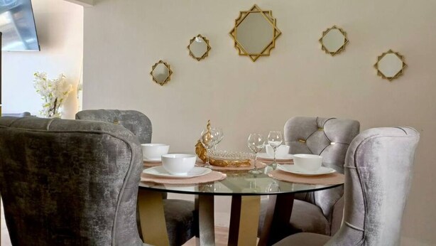 Un coin repas avec une table ronde en verre, des chaises à revêtement gris, de l’assiette blanche et des miroirs décoratifs dorés accrochés au mur.