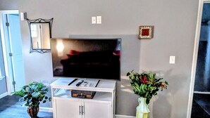 Una sala de estar con una televisión de pantalla plana sobre un mueble blanco, una planta en maceta y un espejo grande en la pared.