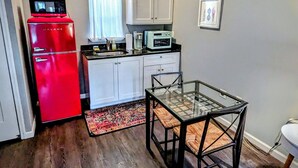 Una cocina compacta con un refrigerador rojo, gabinetes blancos, un microondas y un horno pequeño. Hay una mesa de vidrio con sillas y una alfombra estampada sobre el piso de madera.
