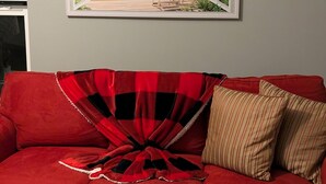 Un sofá rojo con una manta a cuadros rojo y negro y almohadas a rayas, un cuadro de una playa enmarcada en la pared, y una ventana con persianas.