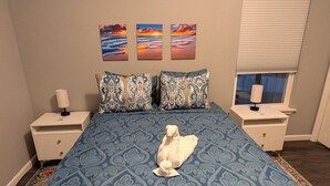 Un dormitorio con una cama, dos mesitas de noche, un ventanal con persianas y tres cuadros en la pared.