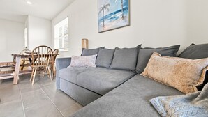 A modern living room with a grey sectional sofa, a wooden dining table with chairs, and a painting of a beach scene on the wall.