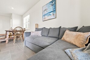 Un salon moderne avec un canapé d’angle gris, une table à manger en bois avec des chaises et un tableau représentant une scène de plage accroché au mur.