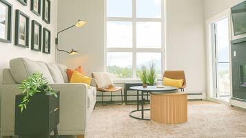 A modern living room with a grey sofa, a round wooden coffee table, and a small round table with chairs. There are framed pictures on the wall, a floor lamp, and a large window providing a view of the outdoors.