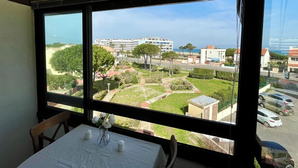 A room with a large window overlooking a landscaped garden, a dining table with a vase of flowers, and a view of a coastal area with buildings and a beach. - 2-Bedroom Apartment, Ocean View, Beach 50 m Away, Air Conditioning, 2 Private Parking Spaces (Saint-Cyprien)