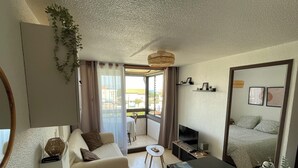 A modern living room with a sofa, a small table, a TV, and a balcony with curtains. - 2-Bedroom Apartment, Ocean View, Beach 50 m Away, Air Conditioning, 2 Private Parking Spaces (Saint-Cyprien)