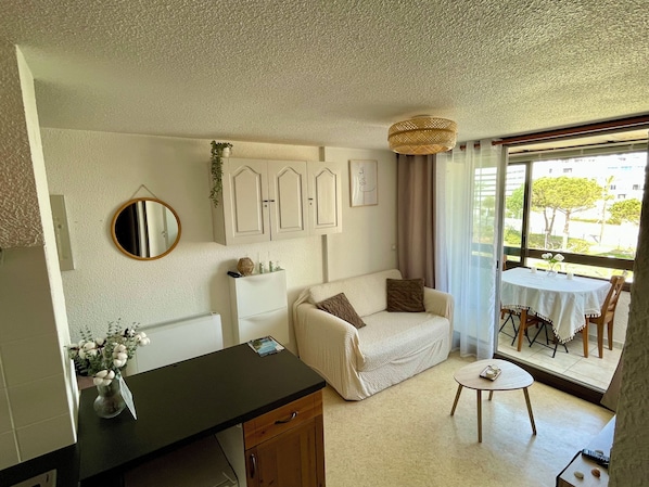 A cozy living room with a sofa, a small round table, a coffee table, a mirror, and a view of the outdoors through a large window.