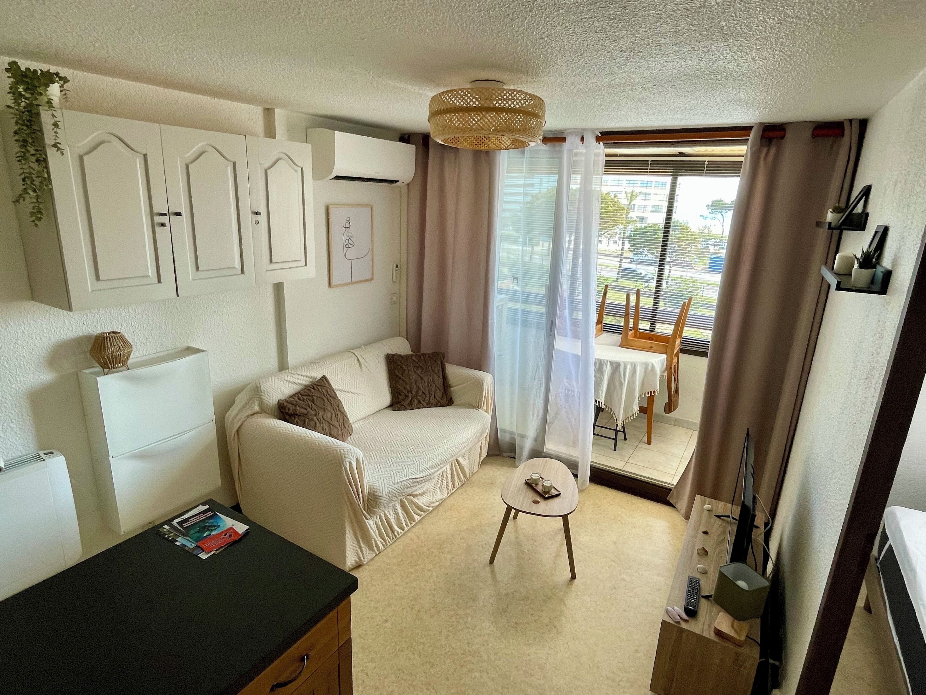 A cozy living room with a sofa, a small round table, and a TV. There is a window with curtains, a plant, and a wall-mounted air conditioner.