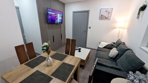 A modern living room with a dining area, a grey sofa, a TV mounted on the wall, and a small table with a vase of flowers. - Spacious and Comfortable Modern Rooms (Clayton)