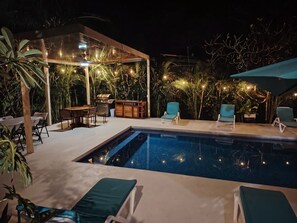 An outdoor pool area with lounge chairs, a barbecue grill, and ambient lighting.