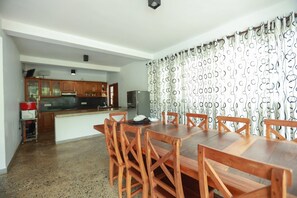 Un coin repas avec une table et des chaises en bois, une cuisine en arrière-plan et des rideaux à motifs.