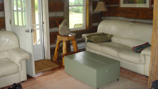 Una habitación rústica de cabaña de troncos con un sofá de cuero blanco, un taburete de madera pequeño y un aparador verde.