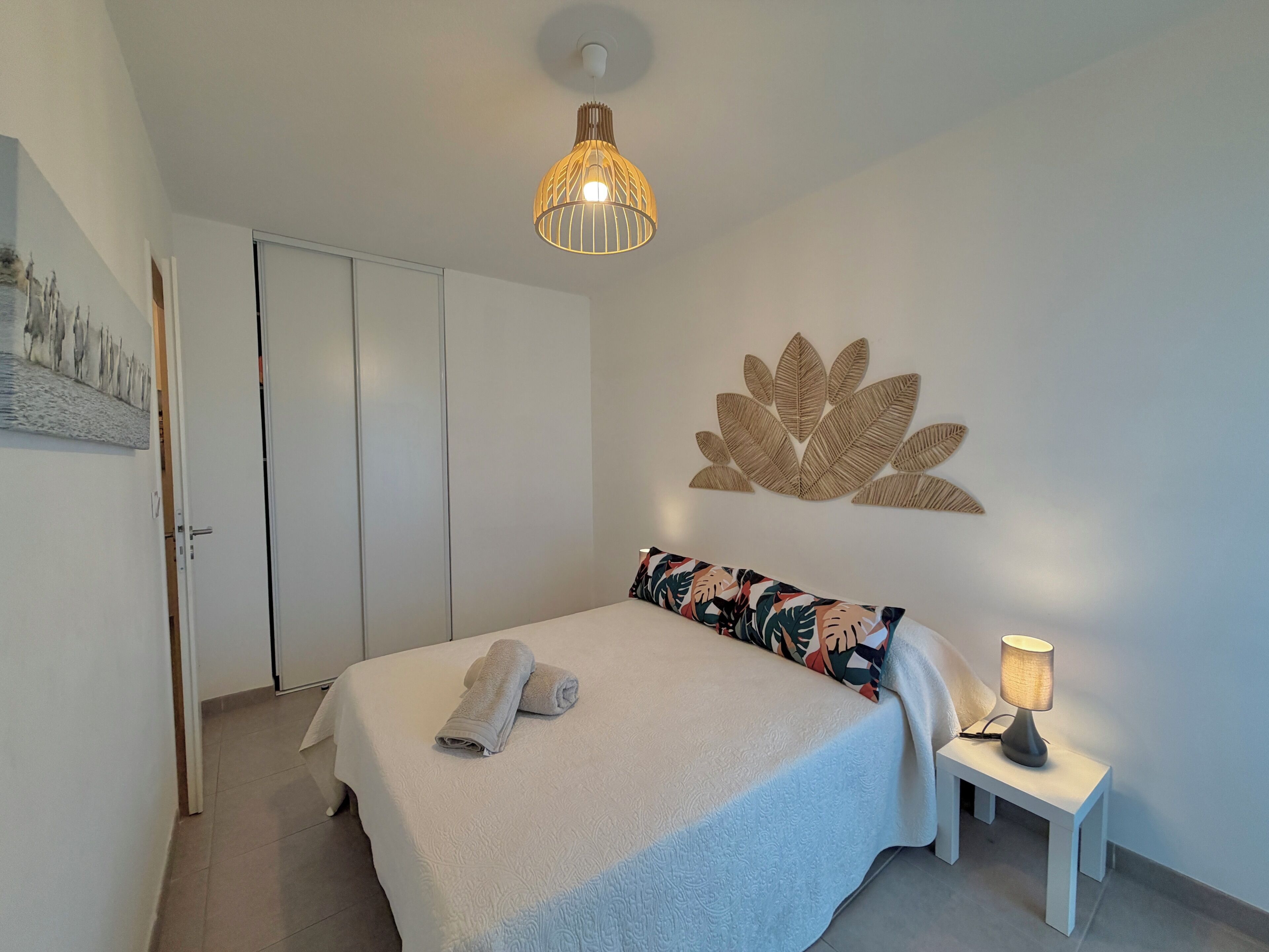 Een net opgemaakt bed met een opgerolde handdoek, een bedlampje en een kunstwerk aan de muur.