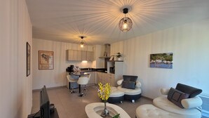 A modern living room with a sofa, a coffee table, and a TV. There is a kitchen in the background with cabinets and a dining area. - Apartment with parking and balcony 200 m from the center (Avignon)