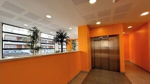 A hallway with orange walls, a tiled floor, and an elevator. There are potted plants along the wall and a view of parked cars outside. - Apartment with parking and balcony 200 m from the center (Avignon)