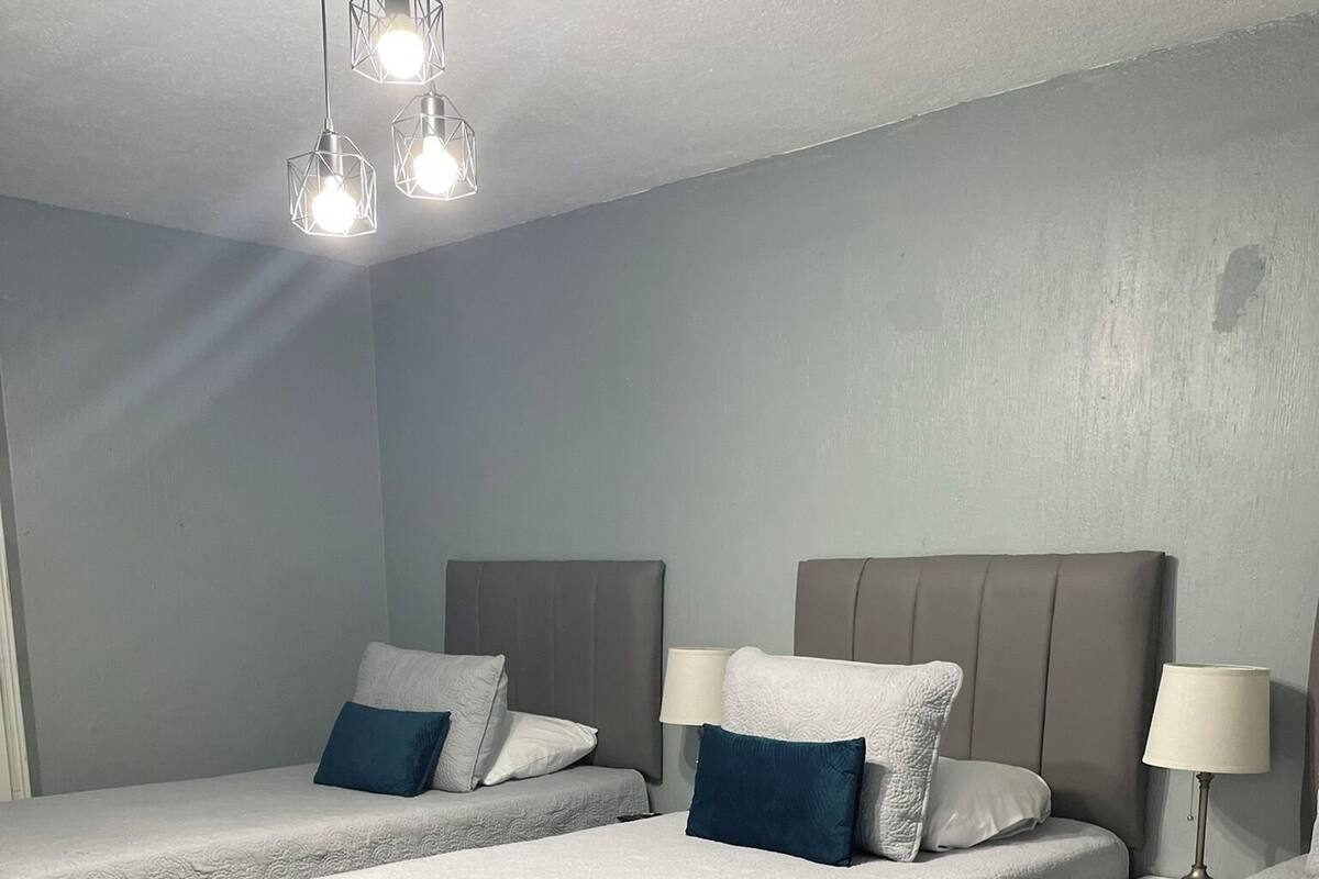 Une chambre d’hôtel avec trois lits, chacun doté d’une tête de lit grise et de linge de lit blanc. On y trouve deux lampes de chevet et un luminaire au plafond.