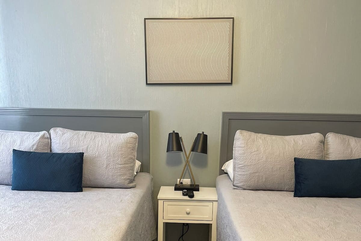 Une chambre double avec deux lits, une table de chevet, une lampe et un tableau encadré au mur.