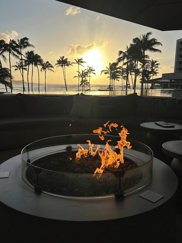 A circular fire feature with flames, a poolside view with palm trees, and a building in the background.