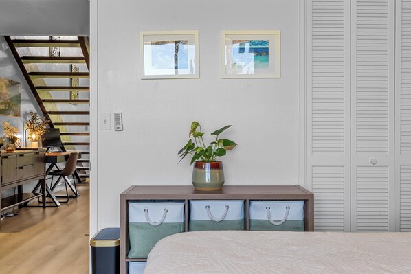 A bedroom with a bed, a desk with a chair, a plant on a dresser, and a staircase in the background.