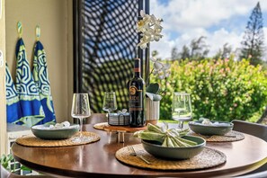 A dining table set with wine glasses, a bottle of Merlot, and bowls of food, with a view of greenery and a patterned wall in the background.