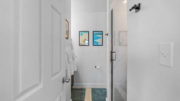 A narrow hallway with white walls, a wooden floor, and a glass door leading to a bathroom. There are two framed pictures on the wall, a towel hanging on the door, and a toilet paper holder.