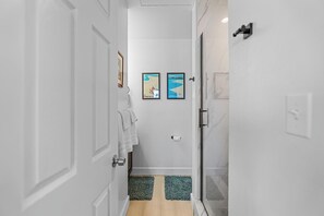 A narrow hallway with white walls, a wooden floor, and a glass door leading to a bathroom. There are two framed pictures on the wall, a towel hanging on the door, and a toilet paper holder. - Turtle Bay Hale: Fully Renovated 2025, Beach-Pool-Golf Course Frontage-A/C (Kahuku)