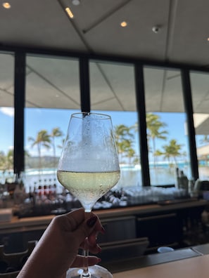 A hand holding a glass of white wine with condensation, a bar area, and large windows with a view of palm trees and a pool.