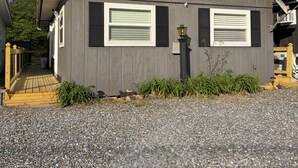 A single-story building with a dark gray exterior, two windows with shutters, a small porch with a wooden railing, and a gravel driveway.