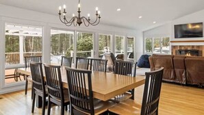 A dining area with a large wooden table and chairs, a chandelier, and large windows offering a view of the outdoors. - Luxury 5b 3ba on 10 acres w Lake view, Firepit, Hot Tub, Gameroom, close to Ski! (Sanbornton)