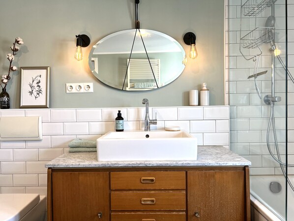 Une salle de bain avec un lavabo blanc, un meuble-lavabo en bois et un miroir au-dessus. On y trouve deux appliques murales et une douche avec une porte en verre.
